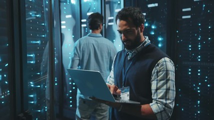 Data center IT administrator working with laptop computer checking rack server cabinets system performance. Webhosting and telecommunication.