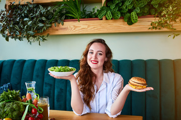Wall Mural - Beautiful young woman decides eating hamburger or fresh salad in kitchen. Cheap junk food vs healthy diet