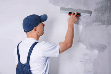 worker in blue overalls and cap plastering a wall with finishing putty using a putty knife. repair w