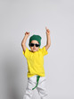 Happy funky child boy holds hands over his head free text copy space. Yellow T-shirt and green hat.