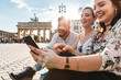 © WILLIAM PERUGINI/Westend61 - Three laughing friends sitting near Brandenburger Tor looking at smartphone, Berlin, Germany
