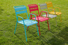 Four Multi-colored Chairs Stand On A Green Lawn On A Summer Evening. Modern Metal Chairs Are Comfortable For Relaxing In An Urban Environment. Sunset Light, Long Shadows