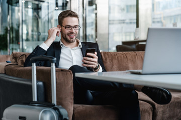 Man with suitcase sitting in airport waiting area while listening music using airpods