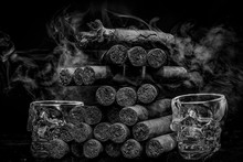 Large Stack Of Hand Rolled Cuban Cigars Against Dark Backround With Smoke And Human Skul Shaped Shot Glasses.