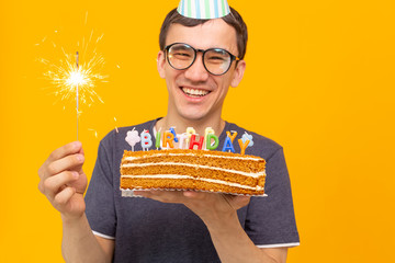 Wall Mural - Positive funny young asian guy with a cap and a burning candle and a homemade cake in his hands posing on a yellow background. Anniversary and birthday concept.