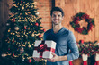 © deagreez - Photo of cheerful positive handsome man smiling toothily holding wrapped gift box present from santa claus standing in front of christmas tree