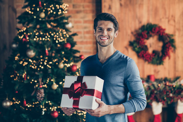 Photo of cheerful positive handsome man smiling toothily holding wrapped gift box present from santa claus standing in front of christmas tree