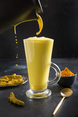 Wall Mural - Pouring glass of ayurvedic golden turmeric latte milk with curcuma powder on black. Vertical shot.