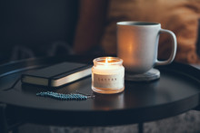 Cozy Interior Ditails. Little Round Table With Cup Of Delicious Tea And Candle, Notepad And Beautiful Blue Color And Silver Earrings. Business Woman Resting After Buzy Day. Selective Focus.