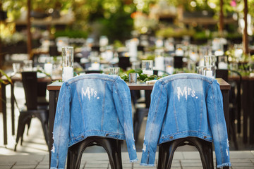 Two bridal blue jeans jackets are hanging on the chairs backs in front of decorated wedding tables