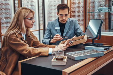 Blonde woman is choosing new pair of glasses while shop assistant is showing her variety of glasses.