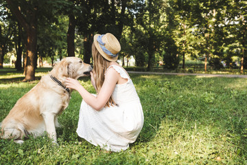 Wall Mural - girl in white dress and straw hat petting golden retriever while sitting on meadow