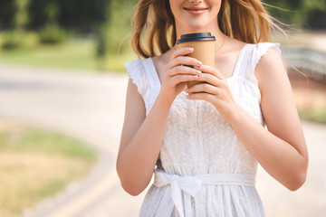 Wall Mural - cropped view of young girl in white dress and straw hat smiling and holding paper coffee cup