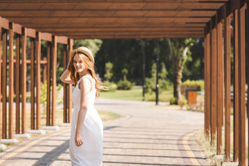 Wall Mural - beautiful girl in white dress touching straw hat while walking near wooden construction and looking at camera