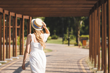 Wall Mural - back view of girl in white dress touching straw hat while walking on pathway near wooden construction