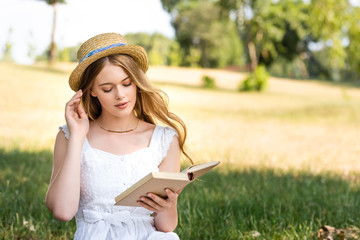 Wall Mural - beautiful girl in white dress touching straw hat and reading book while sitting on meadow