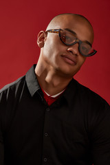 Wall Mural - Close-up shot of a young man with shaved head in a black shirt and with cat eyes retro sunglasses with a brown spotted half-frame and brownish lens. The photo is made on the red background.  