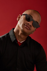 Wall Mural - Close-up shot of a young man with shaved head in a black shirt over a red collar and with cat eyes retro sunglasses with a black half-frame and black lens. The photo is made on the red background.