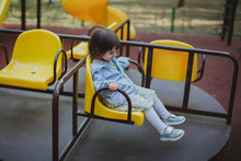 Brunette Girl 2 Years Old In A Dress And Jacket Riding On A Yellow Carousel On The Playground 