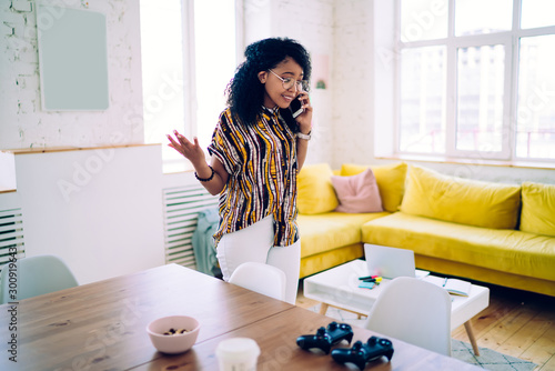 Sad black woman speaking on phone while standing in living room