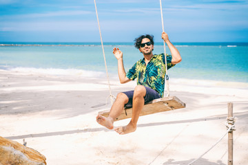 Wall Mural - Stylish happy sexy young brunette man with a beard in a Hawaiian shirt is swinging on a swing on the tropical sea