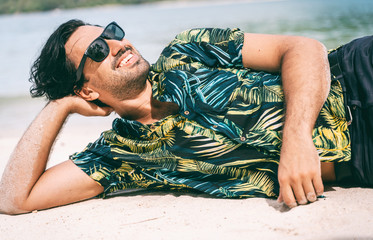 Wall Mural - Stylish sexy young brunette man with a beard in a Hawaiian shirt and sunglasses lies on the sand on a tropical sea beach