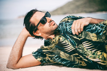 Wall Mural - Stylish sexy young brunette man with a beard in a Hawaiian shirt and sunglasses lies on the sand on a tropical sea beach