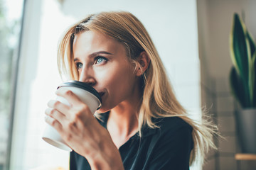 Contemplative hipster girl with blonde hair thoughtful looking away while drinking tasty caffeine beverage from takeaway cup, attractive pensive woman holding coffee and enjoying good taste