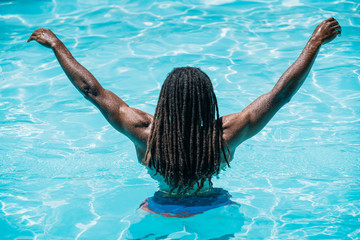 Black boy with dreadlocks on his back in a pool with arms raised