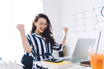 Happy young woman sitting at table with hands up in winner gesture