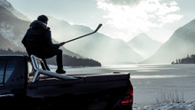  Man Sitting On The Hood Of A Car. Hockey Player Looking For Frozen Pond. Shiny Red Pickup Truck On Ice Covered Road And Snowy Rural Landscape. Offroad 4x4