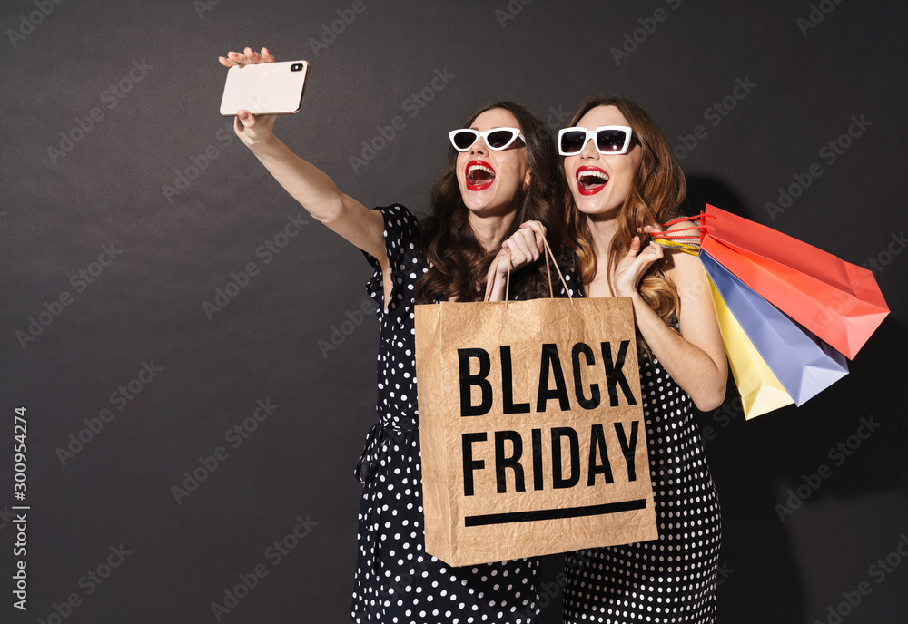 Image of girls taking selfie on cellphones and holding black friday bag - obrazy, fototapety, plakaty 