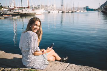 Portrait of positive hipster girl dressed in casual wear smiling at camera while resting in port with sailboats, happy woman tourist with cellphone in hands enjoying time near mediterranean sea