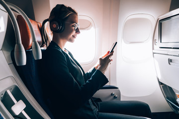 Wall Mural - Smiling female passenger in bluetooth headphones for noise cancellation chatting online via mobile phone using wifi internet connection on board. Young woman in seat near window using smartphone app