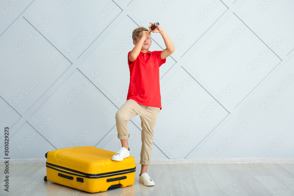 Cute little boy with suitcase and spyglass near light wall