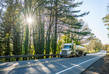 Big rig white day cab semi truck transporting liquid cargo in tank semi trailer running on the forest autumn road