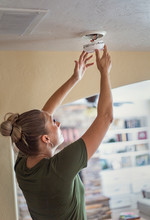 Changing The Battery On A Smoke Alarm In An Interior Of A Home. Woman Standing On A Step Ladder And Fixing The Smoke Alarm.