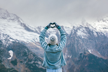 Young traveler girl in gloves standing over mountain peaks and making heart by hands