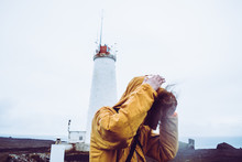 Long-haired Man Wearing Yellow Jacket Struggling Against The Wind While Standing Near Lighthouse In Iceland