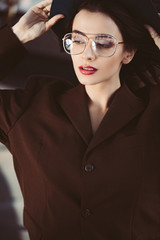 elegant trendy woman posing in hat, eyeglasses and brown jacket on roof