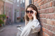 © papa - A happy young woman in sunglasses stands on a city street, leaning against a brick wall.Modern street fashion in the summer season.