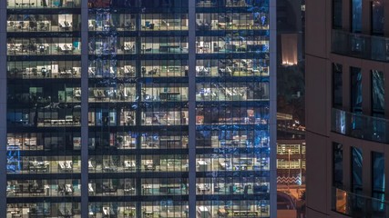 Wall Mural - Office building exterior during late evening with interior lights on and people working inside skyscraper night timelapse. Aerial close up view from above with many illuminated windows.