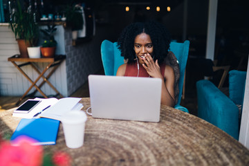 Wall Mural - Amazed dark skinned female blogger feeling excited from received good video review about own web page in social networks using electronic headphones for listening, concept of online multitasking