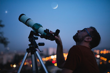 Wall Mural - astronomer with a telescope watching at the stars and moon with blurred city lights in the backgroun