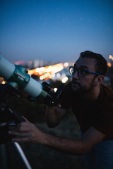 Wall Mural - astronomer with a telescope watching at the stars and moon with blurred city lights in the backgroun