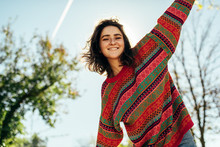 Bottom View Image Of Happy Beautiful Young Woman Smiling Broadly With Windy Hair And Freckles Has Joyful Expression, Wearing Colorful Knitted Sweater With Arms Wide Open, Posing On Nature Sunlight
