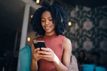 Wall Mural - Positive black hipster girl typing text message for sending to friend connected to wifi internet on cellular phone, happy female teenager playing online games on cellphone during leisure time