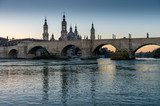 Fototapeta Londyn - View of the medieval stone bridge in front of the Cathedral of El Pilar, Zaragoza (Spain), on the banks of the River Ebro, during the sunset.