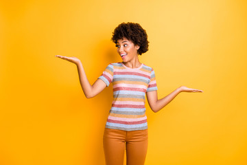 Photo of amazing dark skin curly model lady holding two new products on palms advising cheap one wear casual striped t-shirt pants isolated on yellow background