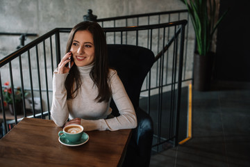 smiling young woman sitting at wooden table with cappuccino and talking on smartphone on balcony in coffee shop